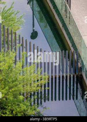 Une scène d'eau sereine avec des poteaux verticaux en bois reflétés dans l'eau calme, entouré d'une végétation luxuriante et d'une structure de verre moderne. Banque D'Images