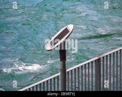 L'invention concerne un luminaire alimenté par énergie solaire monté sur un poteau près d'un plan d'eau, avec une toile de fond d'eau turquoise ondulée et une balustrade métallique. Banque D'Images