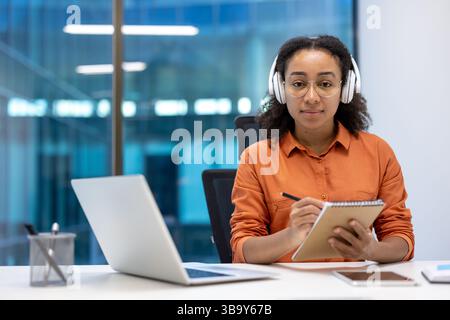 Une femme afro-américaine porte des écouteurs et des lunettes alors qu'elle écrit dans un cahier, prête à commencer son travail. Son ordinateur portable se trouve devant elle. Banque D'Images