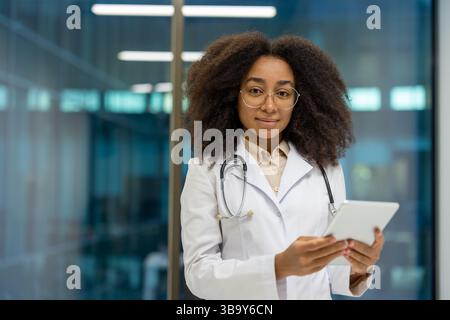Un médecin féminin sympathique tient une tablette dans un bureau moderne, regardant directement la caméra. Elle est habillée d'une blouse de laboratoire et de lunettes. Banque D'Images