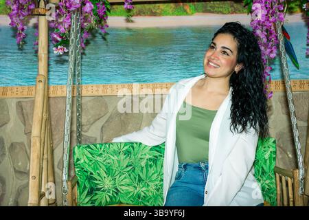 Jeune femme aux cheveux longs, sombres et bouclés souriants assis sur une balançoire décorée de feuilles de cannabis dans un cadre tropical Banque D'Images