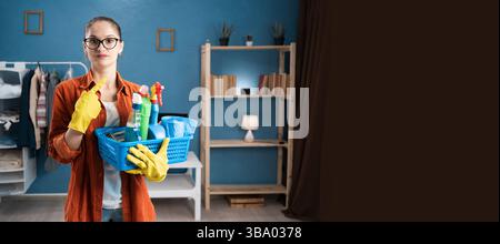 Jeune femme tenant un panier avec des produits de nettoyage debout dans le salon pointant le doigt sur l'espace publicitaire Banque D'Images