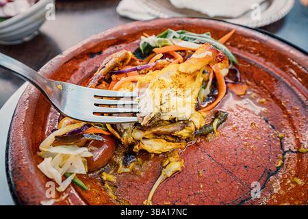Gros plan d'une fourchette ramassant un morceau de poulet d'un tajine marocain traditionnel, servi avec un mélange de légumes colorés dans un plat en terre cuite Banque D'Images