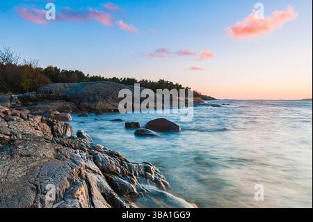 Le rivage de la Suède présente un coucher de soleil magnifique avec des nuages doux se reflétant dans l'eau. Les vagues tapent doucement contre les affleurements rocheux, créant un pois Banque D'Images