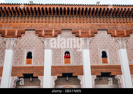 Jeune fille touriste faisant signe d'une fenêtre de la Medersa Ben Youssef, un collège islamique historique à Marrakech, présentant une architecture mauresque complexe Banque D'Images