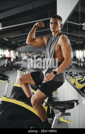 Photo verticale d'un homme sérieux montrant des biceps assis sur la machine à vélo dans une salle de sport moderne spacieuse Banque D'Images