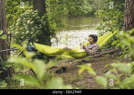 L'homme voyage à vélo, se détendre dans un hamac vert, surfer sur Internet sur un smartphone, écouter de la musique sur des écouteurs dans la forêt près du lac. Cycliste en hammo Banque D'Images