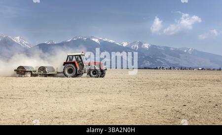 Tracteur tirant un rouleau métallique lourd sur un champ sec sur une belle journée de printemps avec des montagnes en arrière-plan. Préparation du sol, Liptovsky Hradok, Slovaquie, EUR Banque D'Images