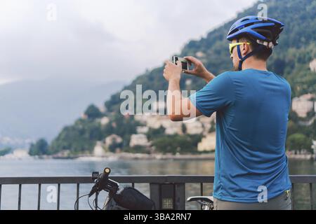 Cycliste prenant des photos avec un téléphone intelligent. Homme caucasien dans le casque de vélo prenant la photo du paysage. Activité en plein air. Touriste fait une photo du lac de Côme i. Banque D'Images