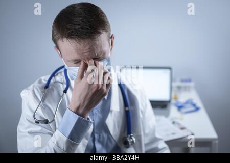 Jeune médecin européen allemand regardant l'appareil photo portant des lunettes et un masque dans le bureau de la clinique sur le lieu de travail avec le regard fatigué. Le sujet est le dur travail de medic Banque D'Images