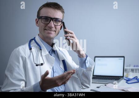 Un médecin européen, portant un manteau médical blanc et des lunettes, consulte le patient sur un téléphone cellulaire dans le bureau de la clinique, assis à une table avec un ordinateur portable. Allemand général Banque D'Images