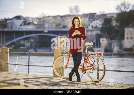 Tourisme thématique sur le vélo et la technologie moderne. Belle jeune femme caucasienne se tient près du vélo rétro rouge sur la rivière Rhin Bâle Suisse victoire Banque D'Images