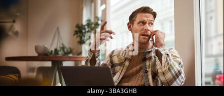 Portrait d'homme d'affaires sérieux parlant au téléphone, assis dans une chaise dans un café avec ordinateur portable, travaillant sur le projet, discutant des questions liées au travail sur Banque D'Images