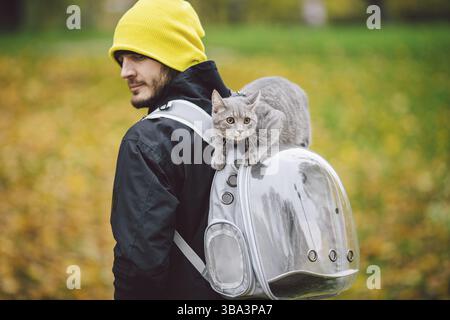 Portrait du grand jeune caucasien heureux homme en plein air jouant avec gris drôle chatons de main assis sur son dos sur transparent chat sacs à dos, Banque D'Images