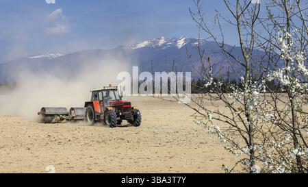 Le tracteur tirant des rouleau métallique, lors de la préparation de terrain sec au printemps, un nuage de poussière derrière, avec des montagnes en arrière-plan Banque D'Images