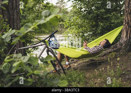 L'homme voyage à vélo, se détendre dans un hamac vert, surfer sur Internet sur un smartphone, écouter de la musique sur des écouteurs dans la forêt près du lac. Cycliste en hammo Banque D'Images