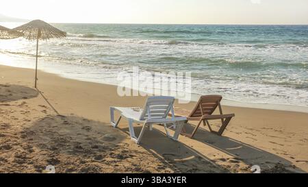 Deux chaises longues en plastique vides face au coucher du soleil, mer en arrière-plan. Image illustrant des vacances tropicales, Lapta, Chypre, Europe Banque D'Images