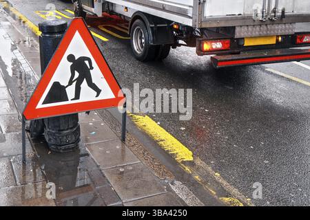 Panneau d'avertissement de travaux routiers à la route humide à côté de la chaussée, fourgon se déplaçant en arrière-plan sur l'arrêt de bus écrivant sur la route, Londres, Royaume-Uni, Europe Banque D'Images