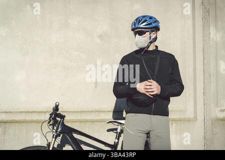 Un cycliste dans un masque protecteur se tient à côté d'un vélo sur un fond de mur gris en béton. Courier inreconnaissable dans le respirateur. Livraison Banque D'Images