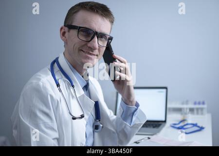 Un médecin européen, portant un manteau médical blanc et des lunettes, consulte le patient sur un téléphone cellulaire dans le bureau de la clinique, assis à une table avec un ordinateur portable. Allemand général Banque D'Images