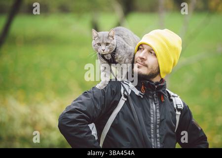 Portrait du grand jeune caucasien heureux homme en plein air jouant avec gris drôle chatons de main assis sur son dos sur transparent chat sacs à dos, Banque D'Images