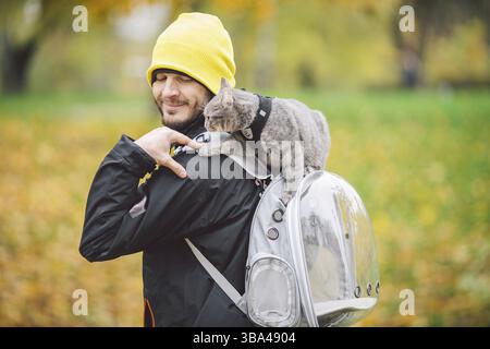 Portrait du grand jeune caucasien heureux homme en plein air jouant avec gris drôle chatons de main assis sur son dos sur transparent chat sacs à dos, Banque D'Images