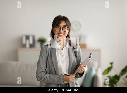 Portrait d'une femme gaie psychologue posant avec un presse-papiers, regardant et souriant à la caméra, travaillant dans un bureau moderne Banque D'Images