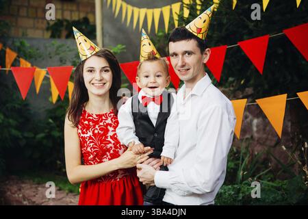 Fête d'anniversaire pour enfants à thème. Père de famille et mère tenant fils d'un an sur fond de verdure et décor festif, guirlandes et colorées Banque D'Images