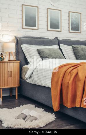 Intérieur élégant de la chambre avec grand lit gris confortable, plaid moutarde en lin et table de chevet en bois près du mur de briques blanches Banque D'Images
