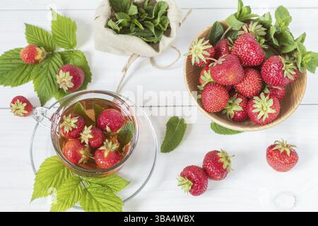 Fraise fraîche dans une assiette en bois. Tasse de verre de thé d'été avec fraise fraîche. Feuilles vertes. Menthe fraîche. Table en bois blanc. Faible profondeur de champ Banque D'Images