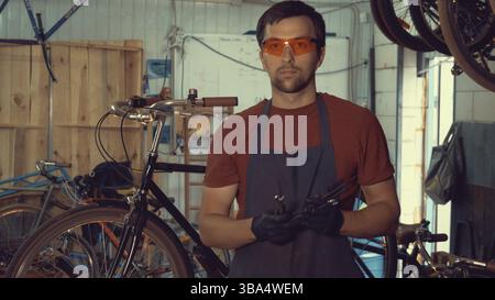 Thème petite entreprise réparation de vélo. Jeune homme brune caucasien dans des lunettes de sécurité, des gants et un tablier posant sur un fond d'un atelier dans les mains Banque D'Images