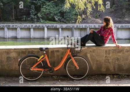 Objet le transport de vélos écologique. Une jeune femme de race blanche dans les jeans et une chemise se trouve l'élève se reposant dans un parc près du lac pour un vélo orange brea Banque D'Images