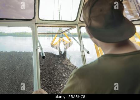 L'opérateur, placé à l'intérieur de la cabine d'une grue flottante, supervise le chargement du charbon sur une barge. Alors que le grand godet de la grue plonge dans un tas Banque D'Images