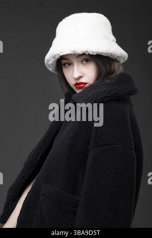 Une belle fille avec un chapeau de fourrure blanc et un manteau de fourrure noir sur un fond gris Banque D'Images