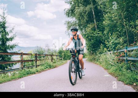 Portrait de femme souriante heureuse habillée de vêtements de cyclisme, casque et lunettes de soleil à vélo sur la piste cyclable asphaltée hors de la ville. Sport actif pe Banque D'Images