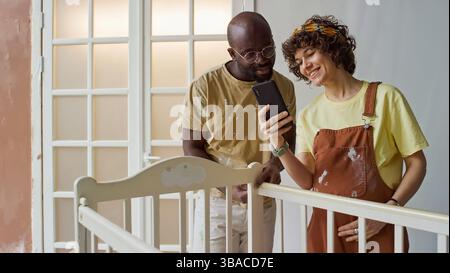 Attendre les parents debout dans la pépinière, regardant smartphone tout en travaillant sur le lit de bébé. Père en chemise jaune et mère en salopette orange, tous deux engagés dans l'activité Banque D'Images