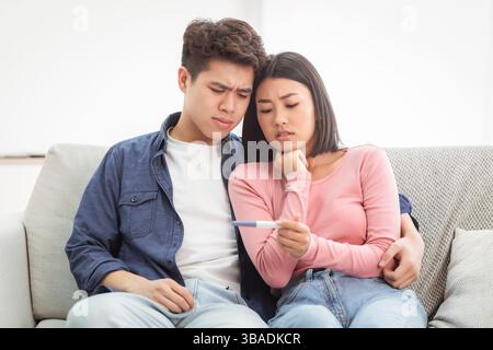 Un couple asiatique frustré qui tient un test de grossesse négatif assis à la maison Banque D'Images