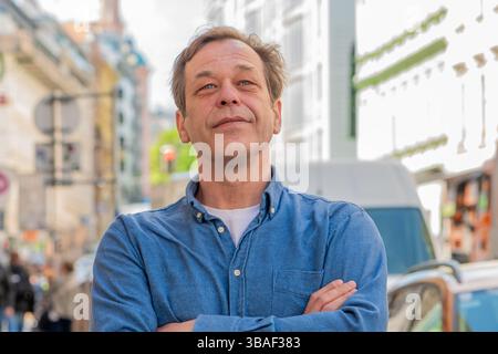 Un homme de 50-55 ans se tient dans une rue avec un arrière-plan flou de piétons et de magasins Banque D'Images