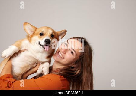 Heureuse jeune femme tenant un chien Corgi mignon souriant, à la fois joyeux, isolé sur fond de studio léger. Banque D'Images