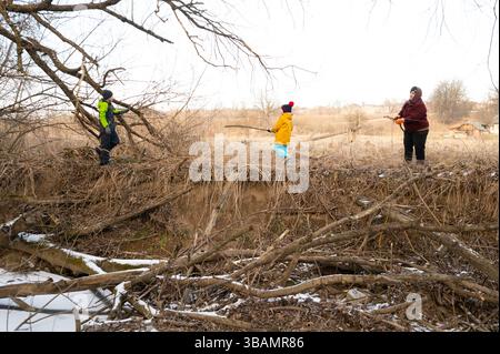Les enfants se livrent joyeusement à des activités ludiques au bord de la rivière, en utilisant des bâtons pour des jeux imaginatifs, entourés par les restes de feuillage hivernal dans un p Banque D'Images