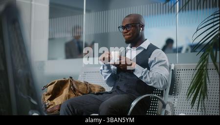 Terminal de l'aéroport international : homme d'affaires afro-américain assis dans le salon des départs, buvant du café. Entrepreneur prospère en attente de vol, en voyage d'affaires. Ralenti, prise de vue moyenne. Banque D'Images