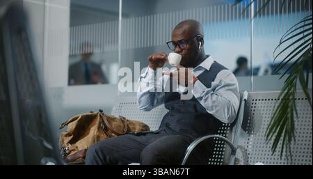 Terminal de l'aéroport international : homme d'affaires afro-américain assis dans le salon des départs, buvant du café. Entrepreneur prospère en attente de vol, en voyage d'affaires. Ralenti, prise de vue moyenne. Banque D'Images
