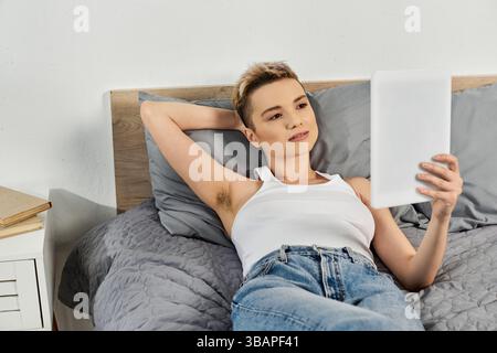 Jeune femme se détend sur un lit, absorbée en lisant une tablette avec un sourire radieux. Banque D'Images