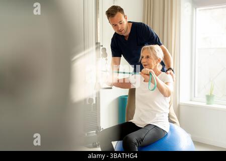 Femme âgée souriante faisant de la musculation avec une bande de résistance tout en étant assise sur un ballon d'exercice, assisté par un physiothérapeute. Concept d'acti Banque D'Images