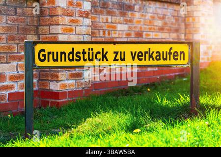 Allemagne - 13 mai 2025 : PHOTOMONTAGE, Un panneau jaune avec l'inscription : propriété à vendre, se dresse sur un espace vert devant un mur et indique une offre immobilière disponible *** FOTOMONTAGE, Ein gelbes Schild mit der Aufschrift : Grundstück zu verkaufen, steht auf einer Grünfläche vor einer Mauer und weist auf ein verfügbares Immobilienangebot hin Banque D'Images