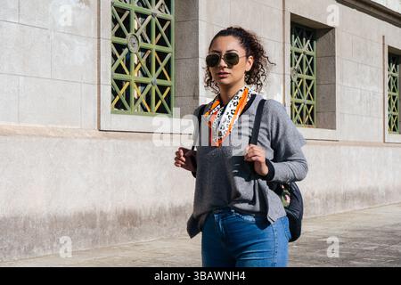 Jeune femme élégante marche près d'un bâtiment, portant un sac à dos et portant une écharpe à la mode Banque D'Images