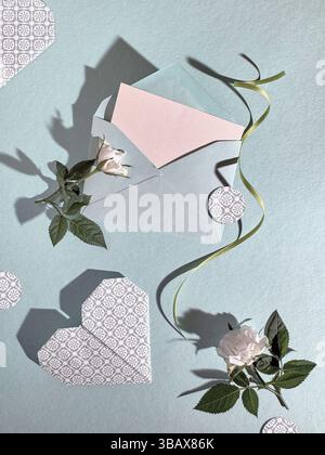 Composition romantique à plat avec enveloppe, carte vierge, roses blanches, découpes en papier en forme de coeur et ruban vert sur fond bleu pastel. Banque D'Images