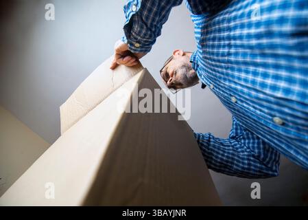 Un homme dans une chemise à carreaux ouvre une grande boîte en carton, vue sous un angle bas. L'image fournit une perspective intéressante soulignant la taille de la boîte Banque D'Images