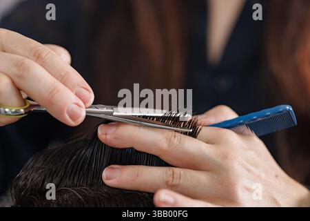Gros plan de styliste coupant les cheveux mouillés avec des ciseaux et peigne pendant la coupe de cheveux détaillée des hommes dans le salon moderne. Banque D'Images