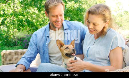 Couple souriant assis sur le siège extérieur dans le jardin d'été avec chien d'animal de compagnie Banque D'Images